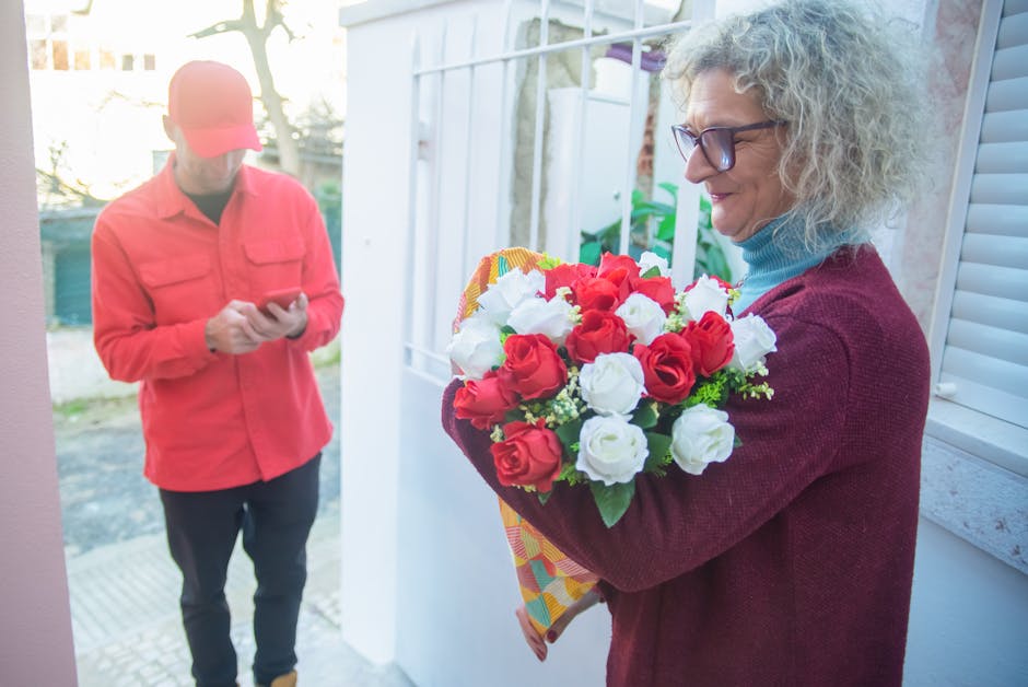 A woman with curly gray hair and glasses, dressed in a maroon sweater and light blue turtleneck, holding a fresh floral bouquet composed of red and white roses, with hints of green foliage, wrapped in