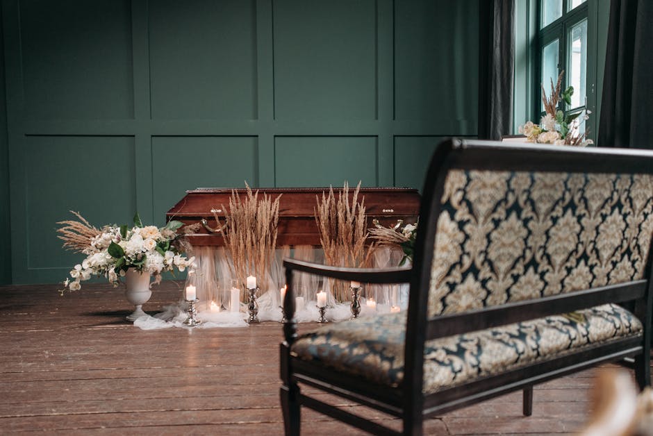 A floral arrangement featuring white roses, peonies, and greenery placed in a white ceramic vase, positioned on a wooden table with soft candlelight surrounding it. Behind the arrangement, a wooden ca
