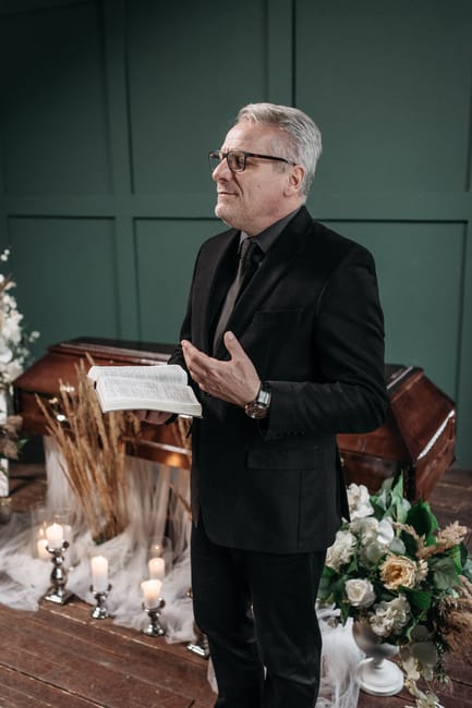 A middle-aged man with gray hair and glasses, dressed in a black suit and dark shirt, stands in a room with dark green paneled walls. He appears to be speaking or giving a speech, holding a booklet or