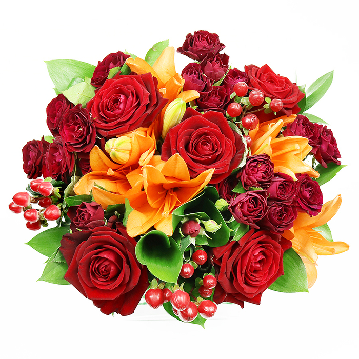 Captured flat against a white background and arranged in a tight, classic round form, this bouquet reads like an autumnal jewel box: deep, comforting reds warmed by sudden flashes of orange and tempered with fresh green. Large, fully blossomed deep-red roses form the heart of the composition; their velvety petals curve inward and reveal the intricate centres that have long made roses synonymous with romance. Bracketed among these are vivid orange Asiatic lilies - unmistakable by their trumpet silhouette and dark-speckled throats - some fully splayed to show their stamens, others half-open or in pointed buds that suggest delicate progression. Accent clusters of small, dark burgundy spray roses fill the negative spaces, their packed rosettes creating depth and a luxurious, layered texture that contrasts with the broader rose blooms. Intermittent clusters of shiny Hypericum berries, in a saturated ruby red, provide glossy, tactile punctuation across the surface. Broad green foliage, likely ruscus or salal, slips in at intervals to give the bouquet structural rhythm and a refreshing colour break against the warm palette. The lighting is gentle and diffuse, allowing textures - velvety, satiny, glossy - to be read clearly without heavy shadow, and the imagined scent is a warm blend of rose depth and the spicy-citrus lift of lilies. The overall impression is one of celebratory richness and considered craftsmanship, ideal for anniversaries, heartfelt congratulations, or a warming centrepiece for a home close to the Thames at Blackfriars or a table near nearby Fleet Street.