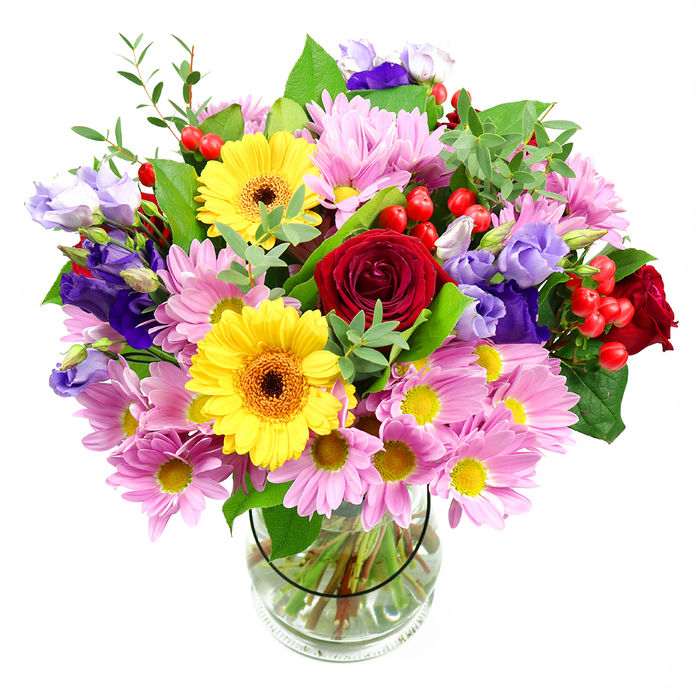 A vibrant mixed bouquet fills a clear glass vase, its rounded shape and generous size creating a strong visual presence against a crisp white background. At the core are sumptuous deep red roses, their velvety petals curling inwards and outwards in intricate layers, giving the arrangement a rich, romantic centre. Flanking them, two bold yellow gerbera daisies beam with sunny confidence, each bloom featuring a dark brown-orange centre that adds both contrast and warmth. Surrounding this centrepiece, abundant pink daisies or chrysanthemums open in a haze of slender petals, their petite yellow centres echoing the gerberas while softening the overall palette. Interwoven with these pastel blooms are purple and lavender lisianthus, their bell-shaped flowers and buds lending vertical accents and cooler tones of violet and lilac that introduce an air of sophistication. Bright red Hypericum berries pop out between the flowers like polished beads, offering small bursts of colour and a rounded texture that contrasts with the flat gerbera faces and feathery chrysanthemum petals. The bouquet is cradled by lush greenery in different shades, from deep, glossy leaves to more delicate light-green stems, which provide structure and a sense of movement. Through the transparent vase, you can see neatly cut stems immersed in clear water, arranged in a careful fan to support the weight of the blooms above. The overall impression is of a joyful yet refined design, suitable for celebrations, congratulations, or simply brightening a workspace near Blackfriars, Fleet Street, or the Thames Path. The styling, with its clean background and even lighting, feels modern and urban, perfectly in tune with the contemporary offices and apartments that line the river here.